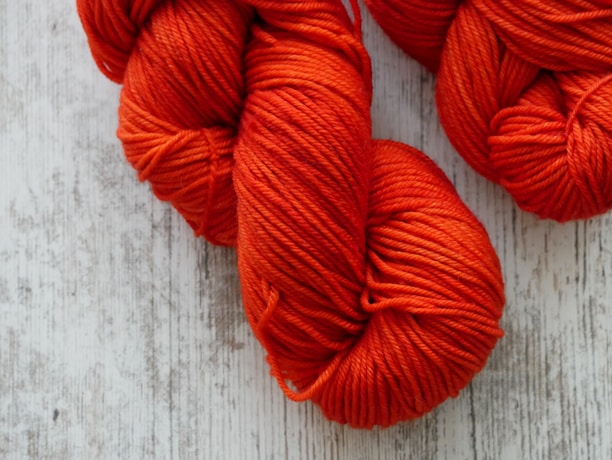 Bundles of bright orange yarn are placed against a textured white wooden surface. The yarn is neatly coiled, exhibiting a thick and uniform texture.