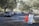 Cars are lined up in a queue on a tree-lined road leading to a white tented area. Orange traffic cones and a sign labeled 'Lane 1' are positioned nearby, guiding the vehicles. The setting appears to be an organized drive-through or testing center.