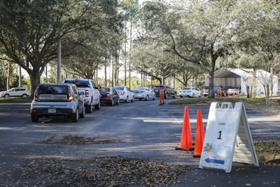 Cars are lined up in a queue on a tree-lined road leading to a white tented area. Orange traffic cones and a sign labeled 'Lane 1' are positioned nearby, guiding the vehicles. The setting appears to be an organized drive-through or testing center.
