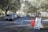 Cars are lined up in a queue on a tree-lined road leading to a white tented area. Orange traffic cones and a sign labeled 'Lane 1' are positioned nearby, guiding the vehicles. The setting appears to be an organized drive-through or testing center.