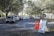 Cars are lined up in a queue on a tree-lined road leading to a white tented area. Orange traffic cones and a sign labeled 'Lane 1' are positioned nearby, guiding the vehicles. The setting appears to be an organized drive-through or testing center.