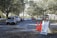 Cars are lined up in a queue on a tree-lined road leading to a white tented area. Orange traffic cones and a sign labeled 'Lane 1' are positioned nearby, guiding the vehicles. The setting appears to be an organized drive-through or testing center.
