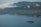 A large, luxurious cruise ship sails across a calm, blue sea with several smaller boats nearby. The coastline features lush green hills and rugged cliffs under a cloudy sky.
