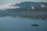 A large, luxurious cruise ship sails across a calm, blue sea with several smaller boats nearby. The coastline features lush green hills and rugged cliffs under a cloudy sky.