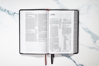 An open book displaying the text of the book of Ezra. It is set on a marble surface with a black cover and white pages. Red and black ribbons are attached to the binding.