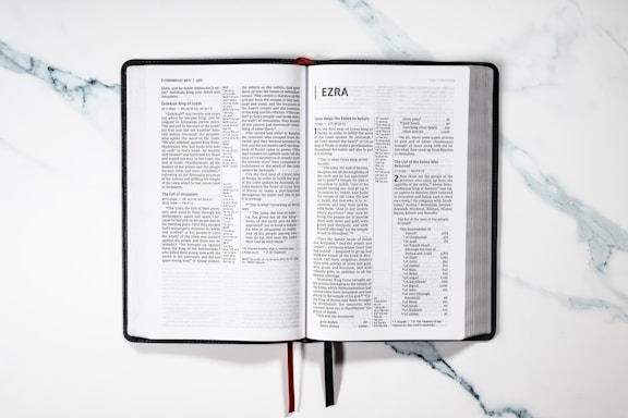 An open book displaying the text of the book of Ezra. It is set on a marble surface with a black cover and white pages. Red and black ribbons are attached to the binding.