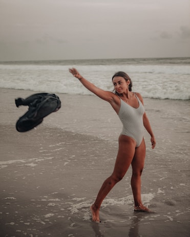 A person in a striped swimsuit is playfully tossing a black garment into the air while standing on a wet, sandy beach. The ocean waves are gently rolling in the background under an overcast sky.