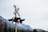 A railway crossing signal with a red and white cross is positioned in front of a backdrop of snow-capped mountains and evergreen trees.