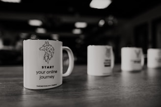 A close-up view of a coffee mug placed on a wooden surface. The mug features a geometric wolf design along with the text 'START your online journey'. The background shows more mugs, slightly out of focus, indicating a row or collection.