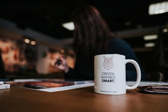 A white mug with an owl illustration and the words 'Driven. Inspired. Smart.' sits on a wooden table. In the background, a blurred individual is engaged in an activity, possibly drawing or writing. The setting appears to be indoors with soft overhead lighting and some art on the walls.
