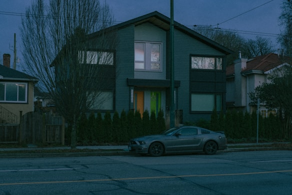 A two-story modern house with a dark facade and large windows is situated along a quiet residential street. A car with a sleek design is parked in front of the house. The area is lined with tall, leafless trees and small shrubs, indicating a season of dormancy. The image is taken during twilight, with a dim, bluish light enveloping the scene.