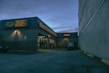 A nighttime view of an automotive repair shop with the name 'Main Street Automotive' illuminated in bright lights. The building has signs for Pennzoil and the entrance is open, revealing vehicles inside the garage. A car is parked in front of the building, and there is a large concrete structure on the right side.