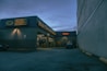 A nighttime view of an automotive repair shop with the name 'Main Street Automotive' illuminated in bright lights. The building has signs for Pennzoil and the entrance is open, revealing vehicles inside the garage. A car is parked in front of the building, and there is a large concrete structure on the right side.