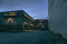 A nighttime view of an automotive repair shop with the name 'Main Street Automotive' illuminated in bright lights. The building has signs for Pennzoil and the entrance is open, revealing vehicles inside the garage. A car is parked in front of the building, and there is a large concrete structure on the right side.
