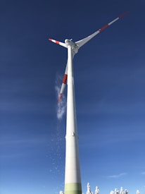 A tall wind turbine stands prominently against a clear blue sky. Its blades are marked with red tips, and snow seems to be falling off them. Snow-covered trees are visible at the base of the turbine, emphasizing a winter setting.
