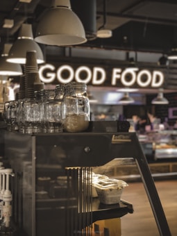 A cozy caf&eacute; interior with a focus on a stack of glass jars and tableware, illuminated by overhead lights. In the background, a glowing sign spells out 'GOOD FOOD', adding a warm atmosphere. The monochromatic ambience is complemented by wood texture and metallic elements, with a hint of natural light.