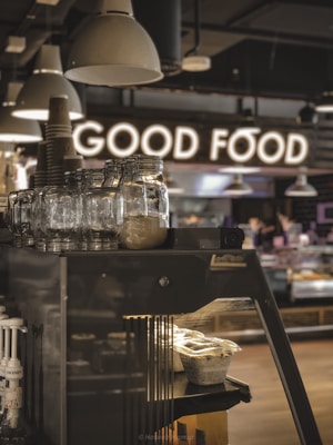 A cozy café interior with a focus on a stack of glass jars and tableware, illuminated by overhead lights. In the background, a glowing sign spells out 'GOOD FOOD', adding a warm atmosphere. The monochromatic ambience is complemented by wood texture and metallic elements, with a hint of natural light.