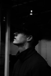 A monochrome profile of a person wearing a cap, looking upwards. The lighting creates deep shadows obscuring much of the facial features and setting a dramatic tone.