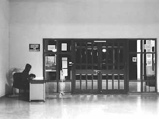 A person sits at a desk with a computer in a mostly empty room, which features a large wooden door with several posted notices and signs. A sign on the wall indicates 'Visitor report to security'. The scene is captured in black and white, giving it a minimalist and somber appearance.