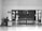 A person sits at a desk with a computer in a mostly empty room, which features a large wooden door with several posted notices and signs. A sign on the wall indicates 'Visitor report to security'. The scene is captured in black and white, giving it a minimalist and somber appearance.
