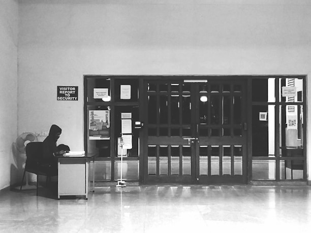 A person sits at a desk with a computer in a mostly empty room, which features a large wooden door with several posted notices and signs. A sign on the wall indicates 'Visitor report to security'. The scene is captured in black and white, giving it a minimalist and somber appearance.
