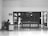 A person sits at a desk with a computer in a mostly empty room, which features a large wooden door with several posted notices and signs. A sign on the wall indicates 'Visitor report to security'. The scene is captured in black and white, giving it a minimalist and somber appearance.