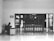A person sits at a desk with a computer in a mostly empty room, which features a large wooden door with several posted notices and signs. A sign on the wall indicates 'Visitor report to security'. The scene is captured in black and white, giving it a minimalist and somber appearance.