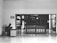 A person sits at a desk with a computer in a mostly empty room, which features a large wooden door with several posted notices and signs. A sign on the wall indicates 'Visitor report to security'. The scene is captured in black and white, giving it a minimalist and somber appearance.