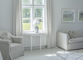 A cozy living room with a soft color palette features a large window allowing natural light to fill the space. There is a comfortable armchair with a patterned cushion, a sleek white side table, and a modern table lamp. Two pieces of framed artwork hang on the wall above a beige sofa with matching cushions.