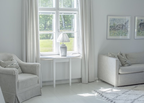 A cozy living room with a soft color palette features a large window allowing natural light to fill the space. There is a comfortable armchair with a patterned cushion, a sleek white side table, and a modern table lamp. Two pieces of framed artwork hang on the wall above a beige sofa with matching cushions.