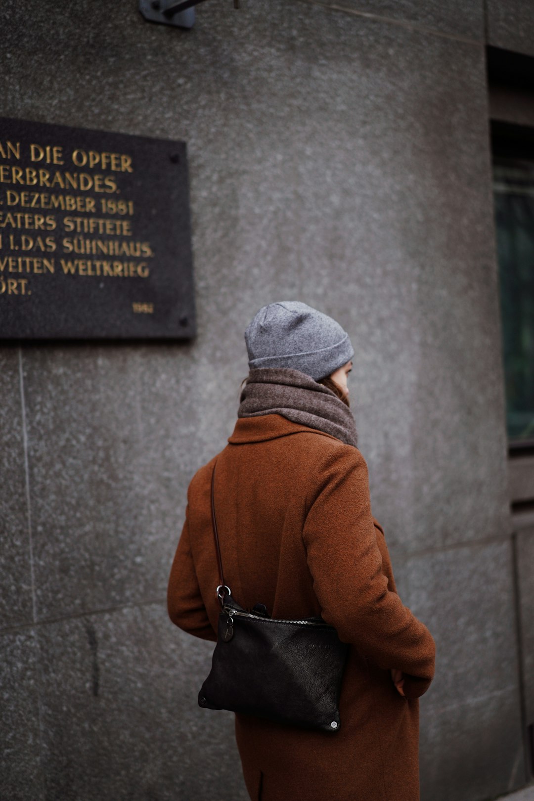 person in brown coat and gray knit cap standing beside gray concrete wall