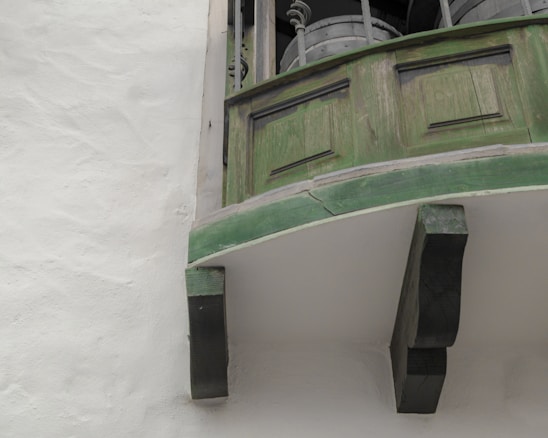 A close-up view of an old wooden balcony with green paint, showing signs of weathering and cracks. The structure is supported by dark wooden brackets, and there are metal railings with cylindrical objects on the floor of the balcony.