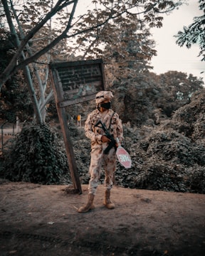 A person in camouflage military uniform stands holding a firearm in one hand and a stop sign with 'Fire' written on it in the other. The background features dense foliage and a weathered, tilted signpost.