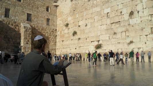 A large stone wall with people standing and praying against it. A seated person is in the foreground, reading or writing on a tablet or book. The stone wall appears ancient and is composed of large blocks with some plants growing between them. The ground is a smooth stone surface.