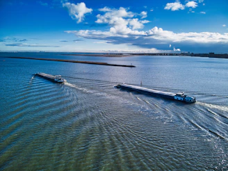 Two long cargo barges travel parallel in a large body of water, creating ripples as they move. The scene includes a vast expanse of blue water and a bright sky with scattered white clouds. The horizon is visible in the distance, suggesting an open and expansive setting.