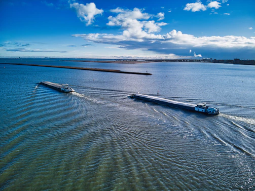 Two long cargo barges travel parallel in a large body of water, creating ripples as they move. The scene includes a vast expanse of blue water and a bright sky with scattered white clouds. The horizon is visible in the distance, suggesting an open and expansive setting.