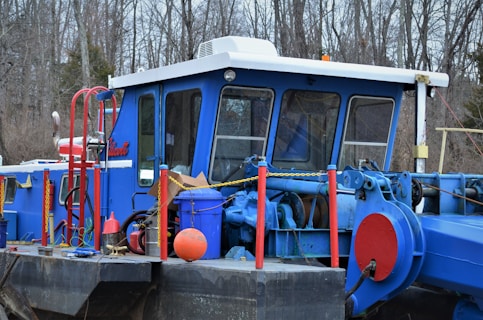 A blue industrial boat or dredger is depicted with a cabin featuring multiple windows and various machinery parts. Red and yellow chains are visible along with safety railings. The scene is situated outdoors, with leafless trees in the background, suggesting a winter or early spring setting.