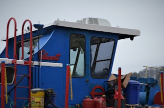 A blue cabin structure of a vehicle or machinery with a sloped roof and large windows. It is surrounded by red railings and various equipment, including a yellow gas canister and other containers. The name 'Ellicott' is visible on the side.