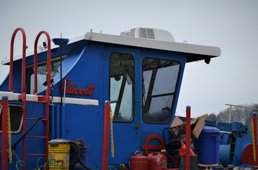 A blue cabin structure of a vehicle or machinery with a sloped roof and large windows. It is surrounded by red railings and various equipment, including a yellow gas canister and other containers. The name 'Ellicott' is visible on the side.