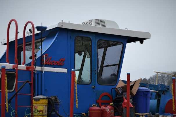 A blue cabin structure of a vehicle or machinery with a sloped roof and large windows. It is surrounded by red railings and various equipment, including a yellow gas canister and other containers. The name 'Ellicott' is visible on the side.