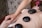 A person is receiving a hot stone massage, lying on a massage table with several smooth, dark stones placed along their bare back. A therapist's hand is visible applying pressure to an arm. A pink flower is tucked behind the person's ear, and the setting appears to be a spa environment.