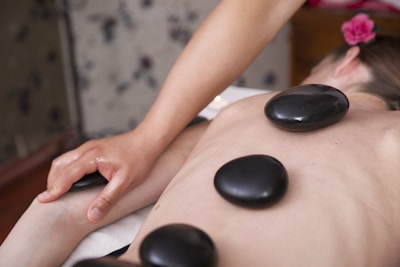 A person is receiving a hot stone massage, lying on a massage table with several smooth, dark stones placed along their bare back. A therapist's hand is visible applying pressure to an arm. A pink flower is tucked behind the person's ear, and the setting appears to be a spa environment.