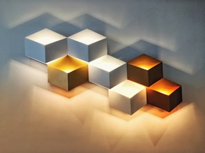 A series of cubic wall lamps arranged in a staggered pattern, each emitting soft light. The cubes vary in color, including white, gold, and copper, creating a warm and modern aesthetic. Shadows cast on the wall enhance the three-dimensional effect.