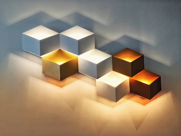 A series of cubic wall lamps arranged in a staggered pattern, each emitting soft light. The cubes vary in color, including white, gold, and copper, creating a warm and modern aesthetic. Shadows cast on the wall enhance the three-dimensional effect.