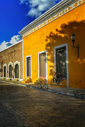 man in black jacket and blue denim jeans sitting on black bicycle near yellow concrete building