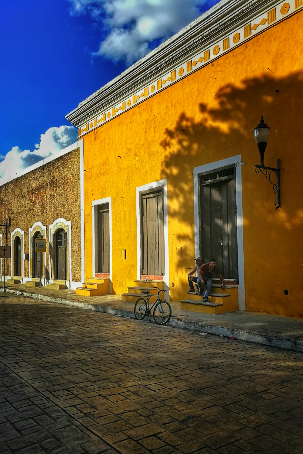 man in black jacket and blue denim jeans sitting on black bicycle near yellow concrete building