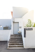 A minimalist modern entrance to a building features a small set of gray stairs leading to a light gray door. A potted plant with white flowers is placed on the top step beside the door. The wall to the right has a sign with the text 'H&ouml;lzelweg' and other smaller text. The architecture is characterized by clean lines and white curved walls, with some greenery visible in the background.