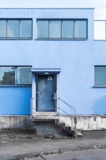 A modern building facade with a light blue exterior and horizontal rectangular windows. The entrance features a metal door marked with the number 24, accessed by a short flight of concrete steps with metal railings. The ground surrounding the building appears slightly overgrown with weeds.