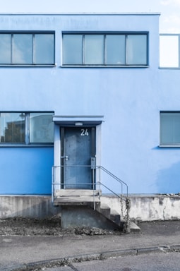 A modern building facade with a light blue exterior and horizontal rectangular windows. The entrance features a metal door marked with the number 24, accessed by a short flight of concrete steps with metal railings. The ground surrounding the building appears slightly overgrown with weeds.
