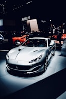 A sleek, silver sports car is prominently displayed under spotlights, surrounded by a crowd in a dimly lit showroom. The background features branding or signage related to the manufacturer.